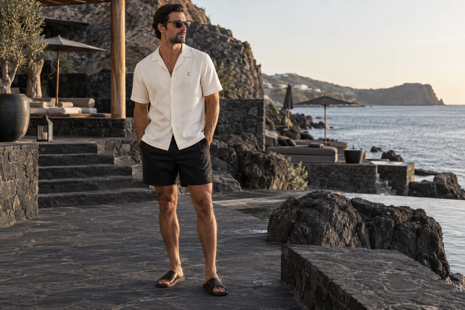 Adult male model in an ivory Cortex relaxed resort shirt and black tailored swim shorts beside a coastal beach club. Original campaign concept.